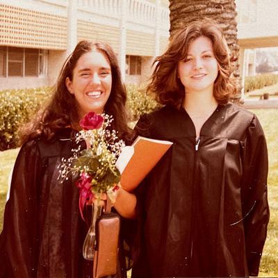 Older photo of alumna Kathy Kustara and friend in graduation gown in front of Seaver Hall